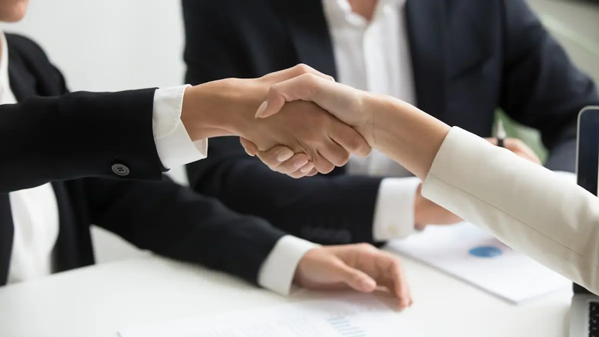 Two people shaking hands across a desk with an open laptop between them.