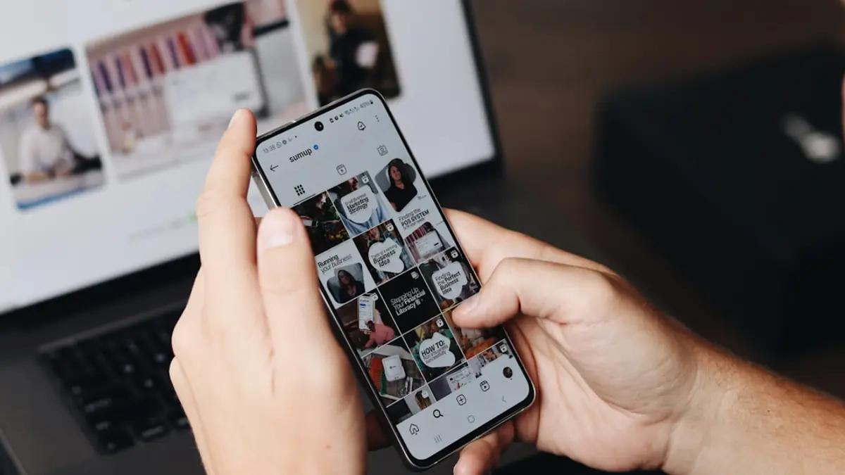 A person holding a phone showing a social media profile, with a laptop behind.
