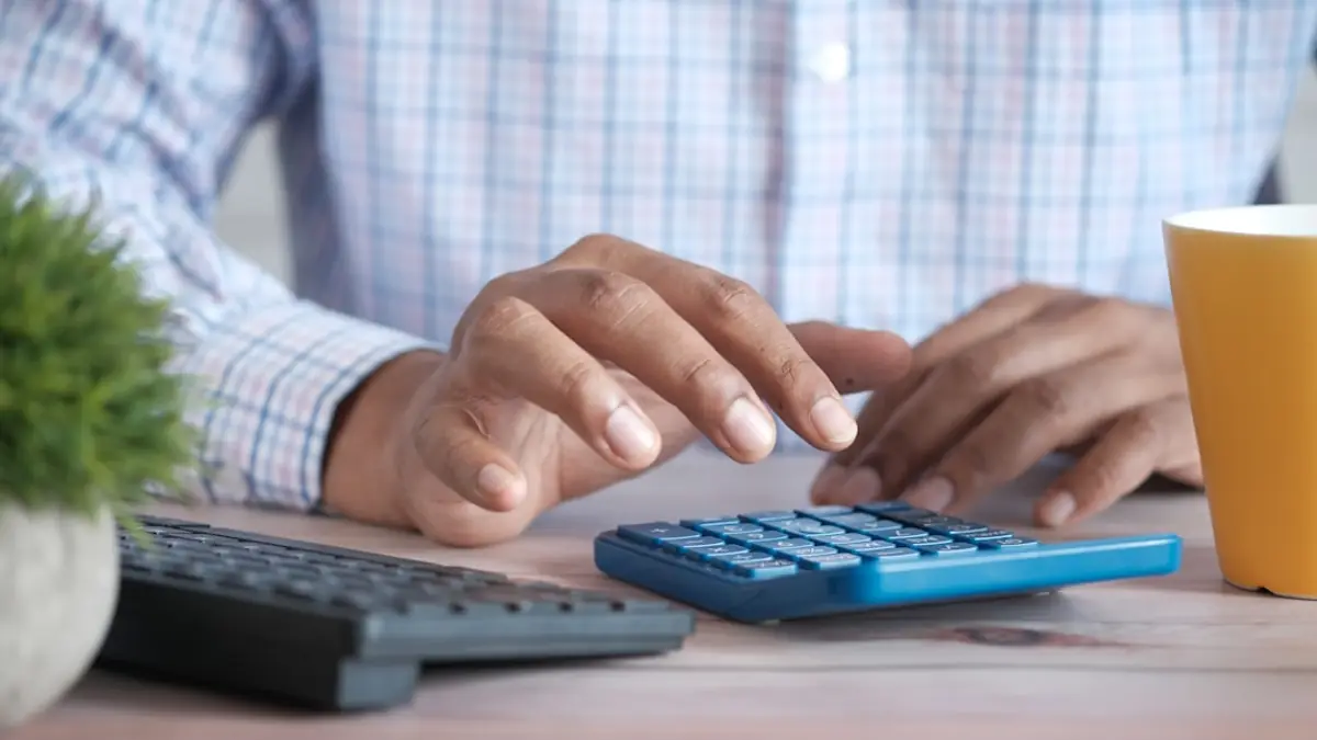 A hand using a desk calculator next to paperwork and a coffee mug.